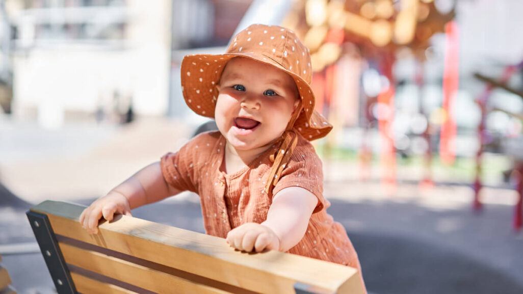 Lachendes Baby mit Sonnenhut, das sich auf eine Bank hochgezogen hat.