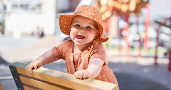 Lachendes Baby mit Sonnenhut, das sich auf eine Bank hochgezogen hat.