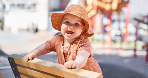 Lachendes Baby mit Sonnenhut, das sich auf eine Bank hochgezogen hat.