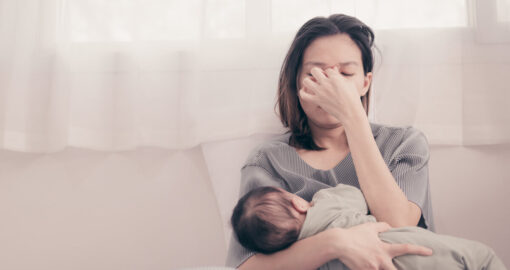 Mutter stillt ihr Baby auf dem Sofa und hält sich erschöpft die Hände an die Stirn.