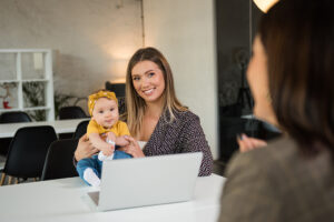Mutter mit Baby sitzt vor einem Laptop beim Gespräch und lächelt strahlend