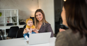 Mutter mit Baby sitzt vor einem Laptop beim Gespräch und lächelt strahlend