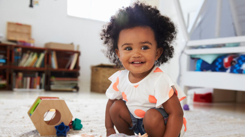 Kleinkind sitzt lächelnd mit Spielzeug auf dem Boden im Kinderzimmer.