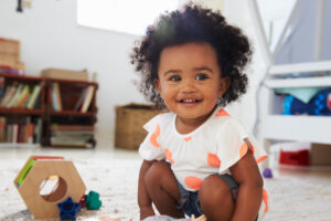 Kleinkind sitzt lächelnd mit Spielzeug auf dem Boden im Kinderzimmer.