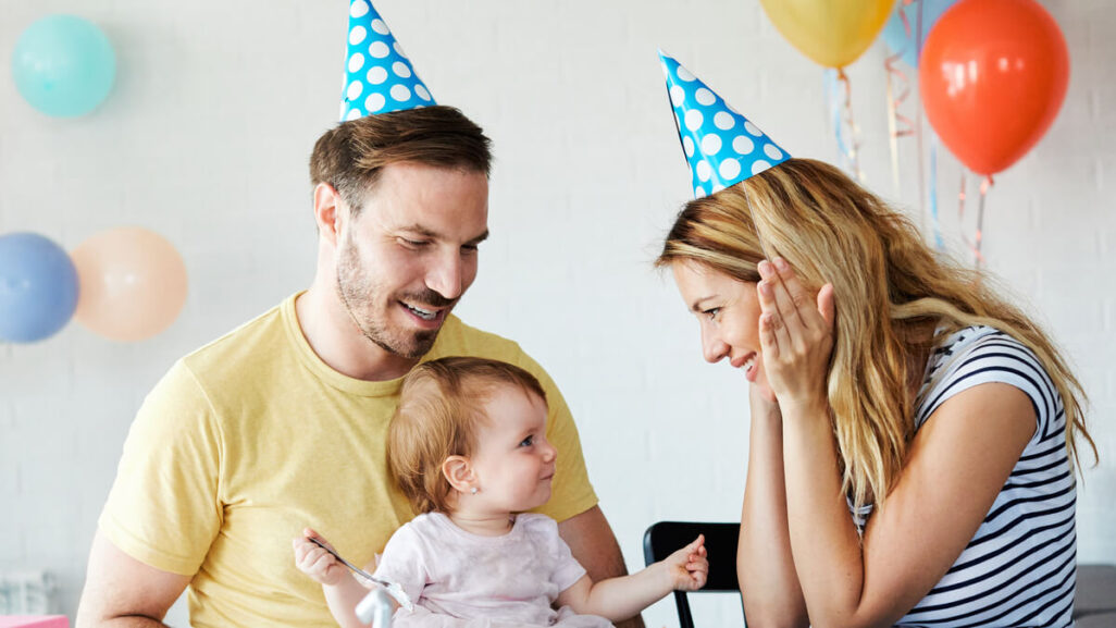 Eltern mit Partyhüten auf dem Kopf feiern zusammen mit dem Baby Geburtstag.