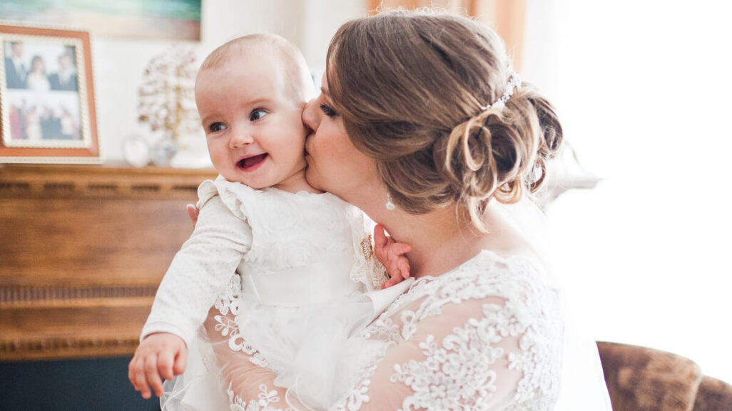 Mutter in Brautkleid küsst ihr Baby auf die Wange.