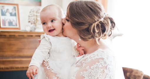 Mutter in Brautkleid küsst ihr Baby auf die Wange.
