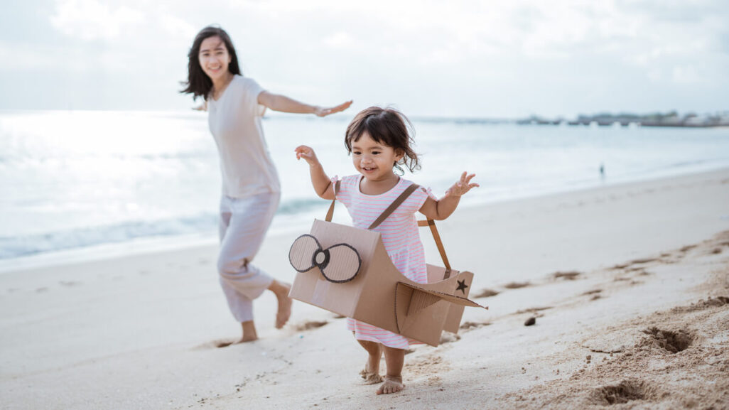 Kind läuft mit Mutter freudig am Strand entlang und trägt dabei ein aus Karton selbstgemachtes Flugzeug um den Körper.