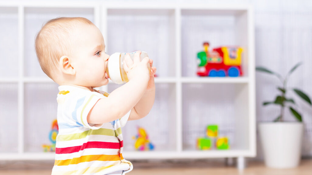 Baby sitzt im Kinderzimmer und trinkt Kamillentee aus einem Fläschchen